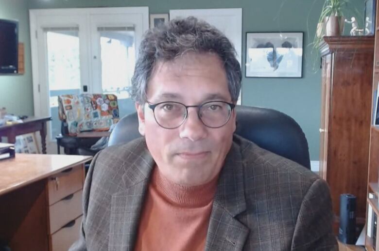 A man with grey hair wearing a checked blazer sits in an office. 