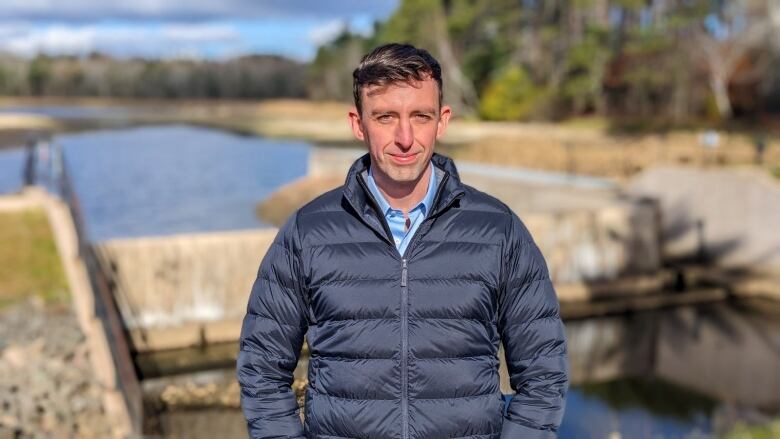 A man in a blue jacket stands in front of a dam 