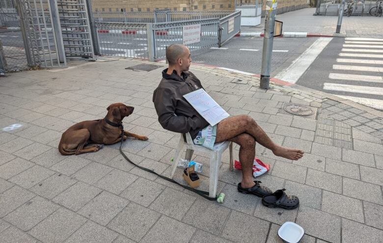 A man sits on a plastic chair on the street, with his dog sitting close by. They are both looking away from the camera.