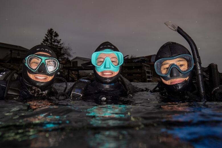 Three people are wearing scuba suits and diving gear. They are smiling at the camera while in the water. 