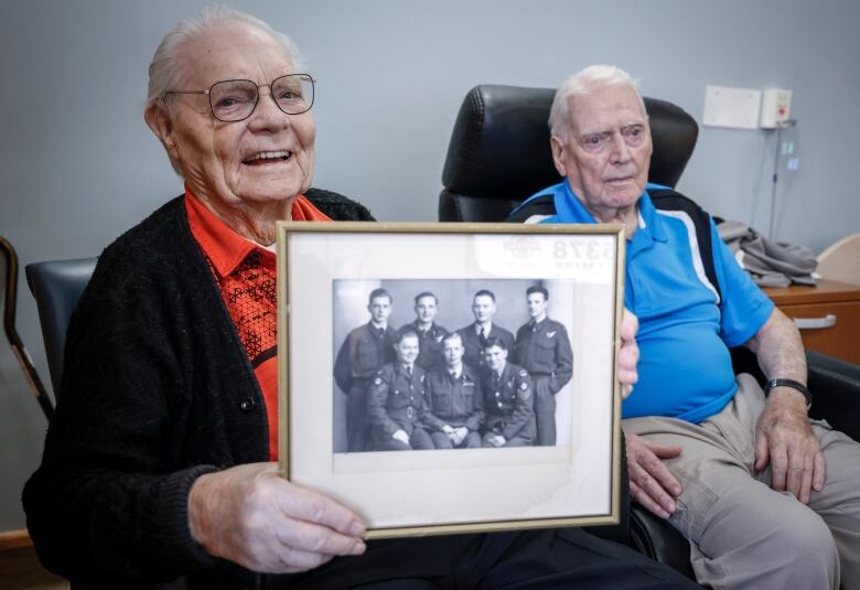 Two elderly men sit. One is holding a photo of a World War 2 bomber crew, to which both men belonged.