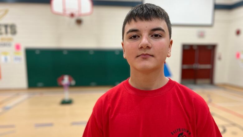 A boy in a red T-shirt looks at the camera as he stands in a school gym.