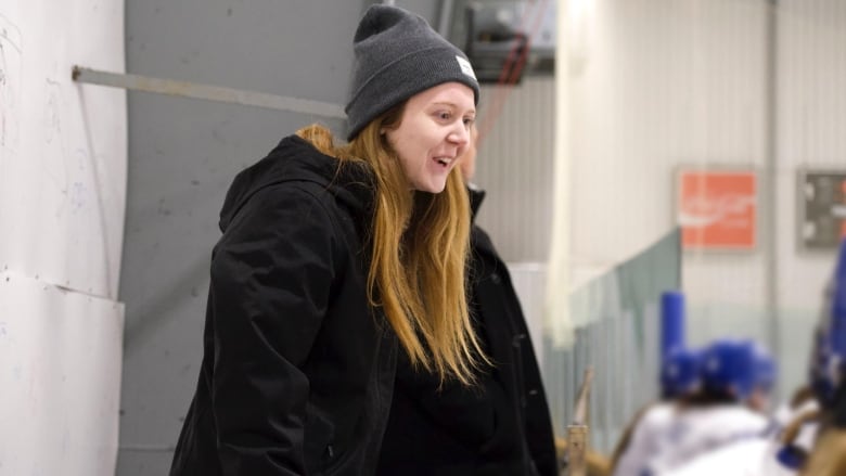 A smiling woman in a tuque stands behind hockey players sitting on a bench.