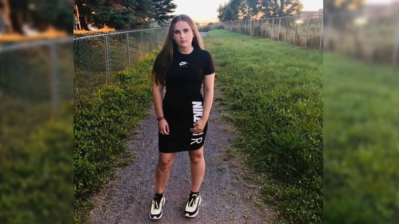 A girl stands on a path surounded by grass and a fence. 