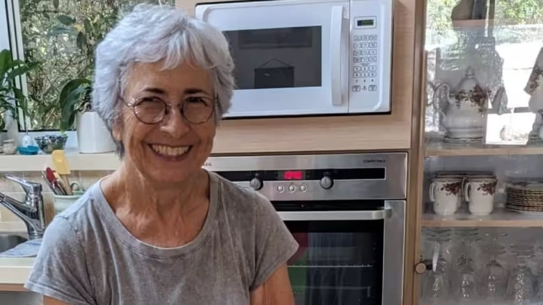 A woman with short white hair and a wide smile looks at the camera. She sits in a kitchen.