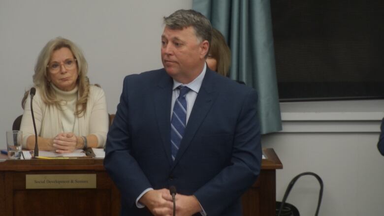 A man in a dark blue suit and tie stands while taking questions from opposition members.