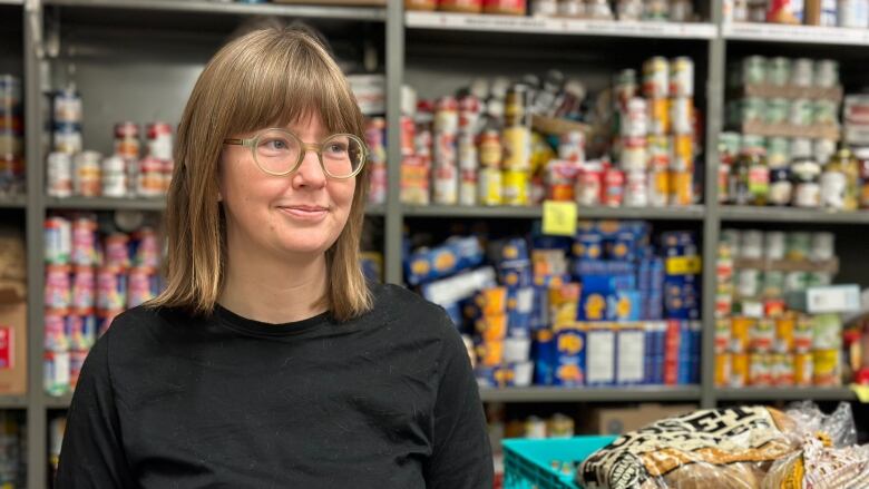 A woman with brown hair and glasses stands in front of shelves full of canned food and boxes.