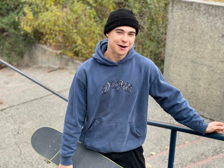 A boy in a hoodie and a beanie hat holding a skateboard and smiling for the camera