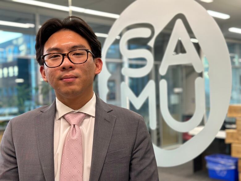 A man wearing a suit and a pink tie stands in front of a glass wall with the letters S-A-M-U written on it.