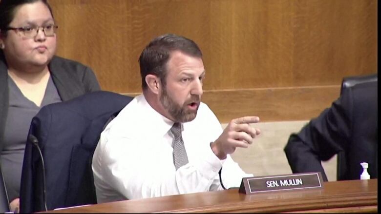 A man sits behind a table, in a white shirt and tie, raising his right hand to point at someone. 