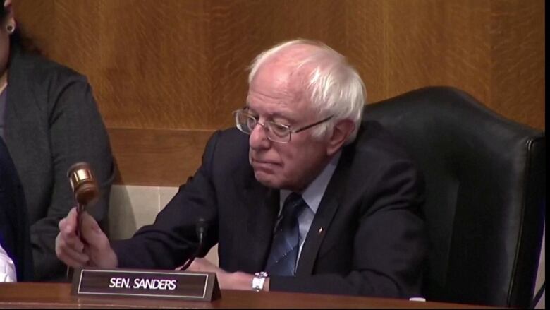 A man with thin, white hair and glasses raises a gavel to hit on the table he is sitting at.