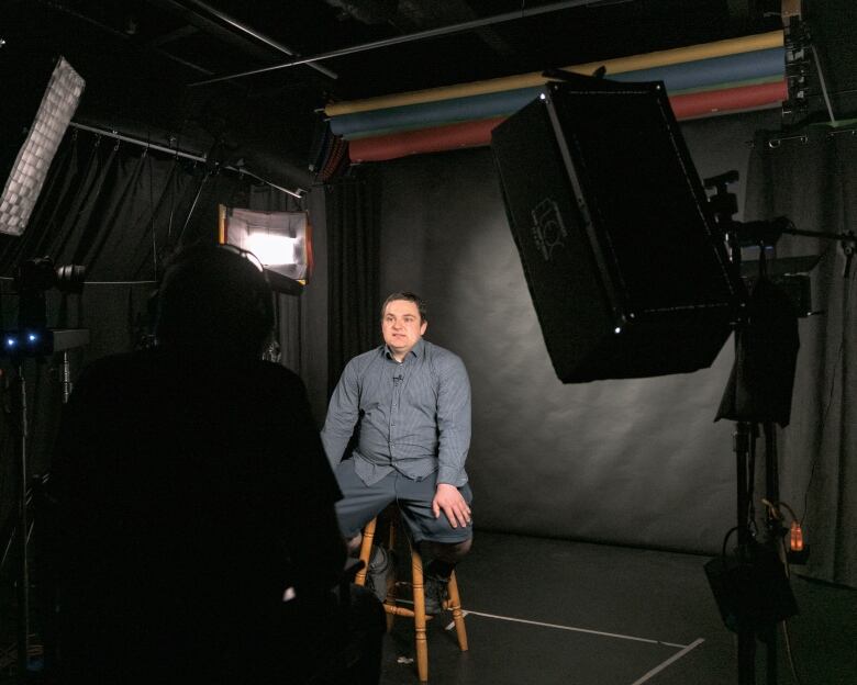 A man wearing grey speaks to a camera while lights and cameras surround him.