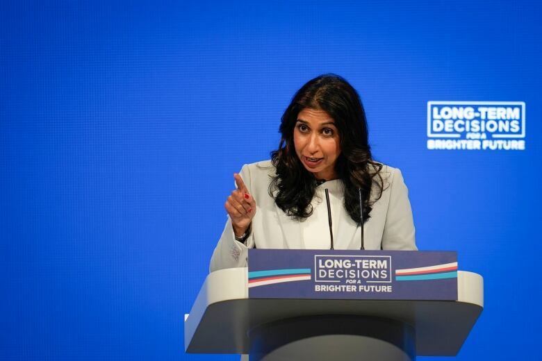 A woman in a blazer points while speaking at a podium at a public event.