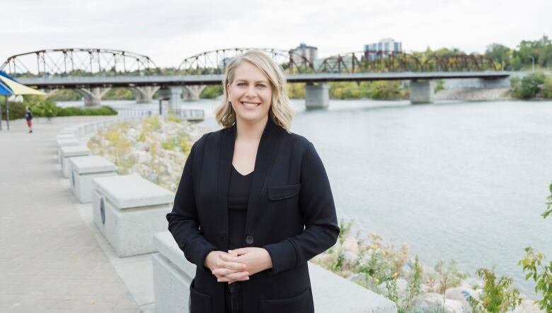 A woman in a black blazer and pants stands in front of a bridge outside