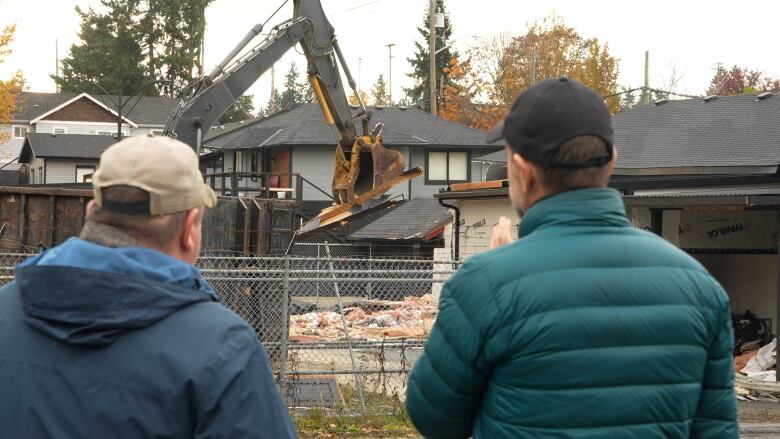 Two men wearing green jackets face away from the camera watching an excavator demolish a building behind a chain-link fence.