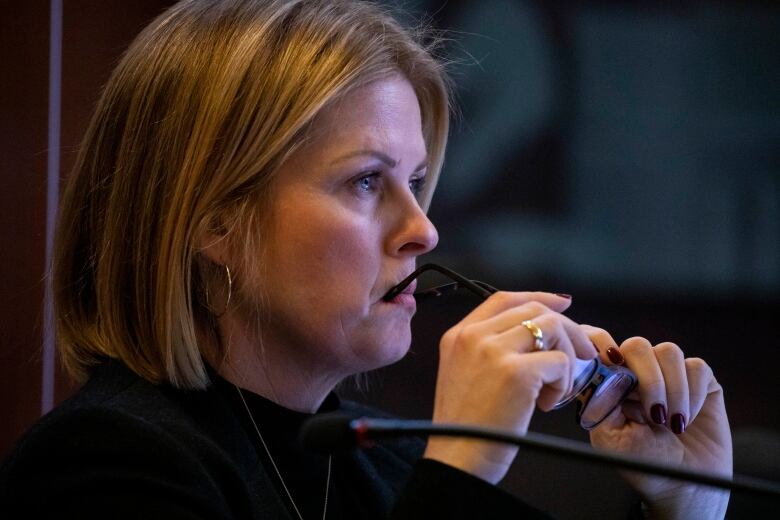 A white woman with short blonde hair places her glasses on her mouth in a pensive motion.