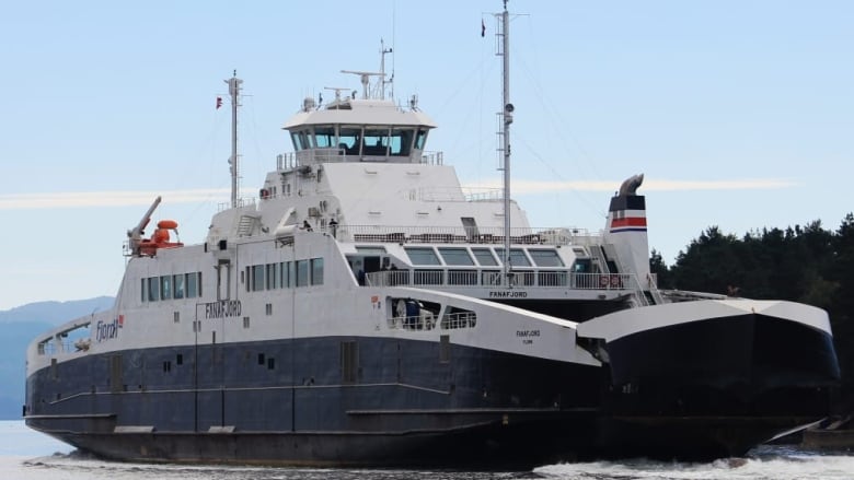 A large ferry with its nose cone opening to let vehicles drive off is shown approaching a dock. 