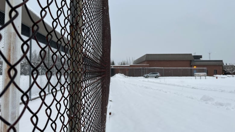 A large brick building is seen in the distance behind a chain link fence. There is snow on the ground.