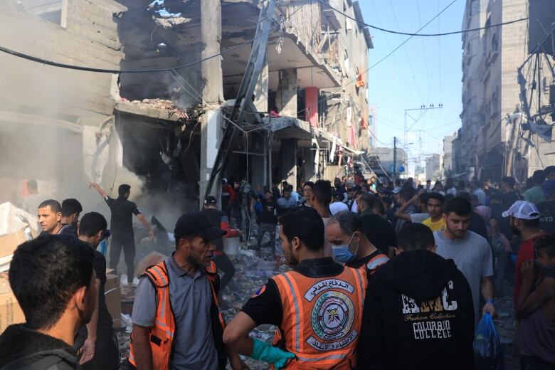 Several people, including some in reflective vests, stand around in an urban area in which debris and damaged concrete buildings are shown.