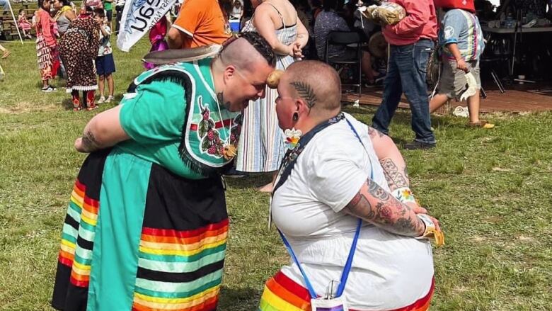 Two people dressed in long skirts with rainbow patterns on them crouch facing each other with a potato between their foreheads.