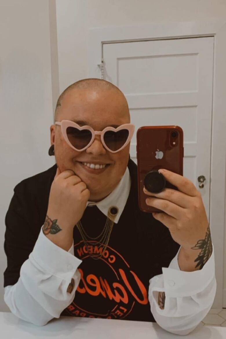 A smiling person with a shaved head and pink heart sunglasses holds up a red phone in front of a mirror.