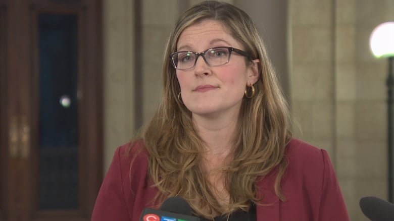 A woman with long brown hair and glasses wears a burgundy blazer. She stands at a podium and speaks to reporters.