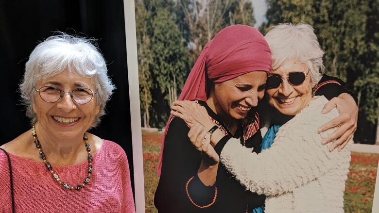 A smiling gray-haired woman stands next to a large photo of herself hugging another woman in a pink headscarf. It's a sunny day and both women are smiling brightly. 