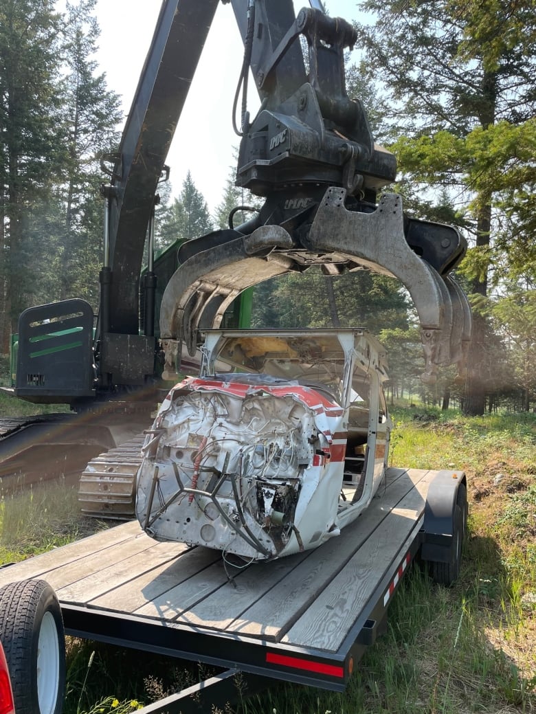 A bashed up plane fuselage is placed on a trailer bed using a digger claw, amid the backdrop of a forest