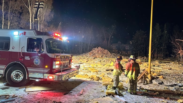 A fire truck with its lights on and 2 firefighters stand in the dark near a pile of building rubble.