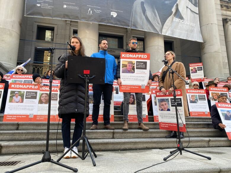 A woman speaks to a mic, flanked by others, in front of a series of placards.