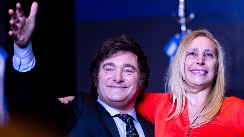 A man in a suit and tie lifts his arm in a celebratory gesture while having his other arm around a woman with shoulder length hair who is smiling.