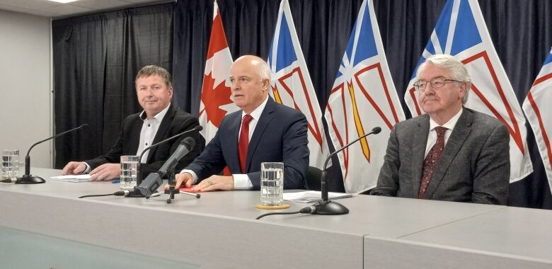 Three men in suits sitting at a table with Canada and Newfoundland and Labrador flags behind them.