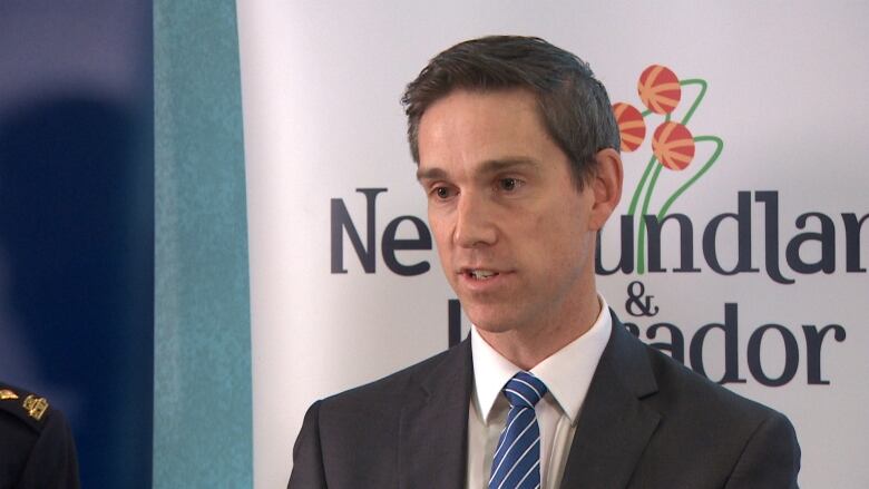 A man wearing a black suit stands in front of a banner with the Newfoundland and Labrador logo on it.