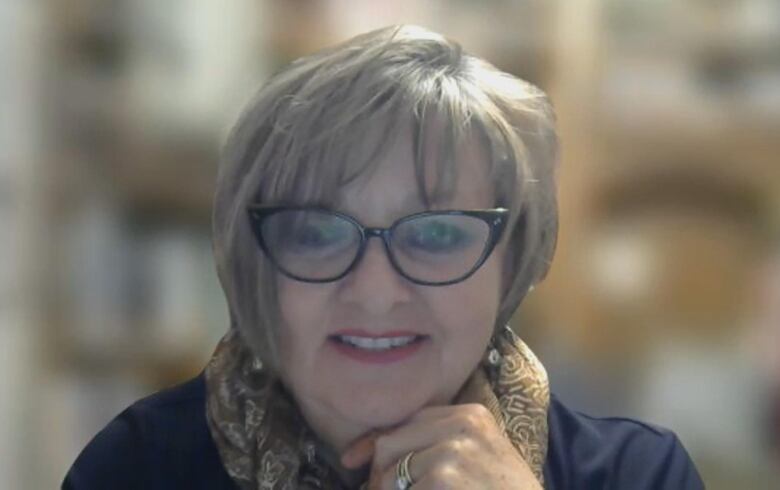 A woman with short hair smiles at her web camera in a Zoom call, wearing a scarf and glasses.