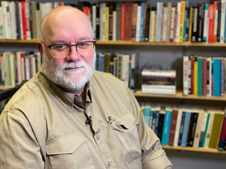 A man wearing a beige shirt sits near a bookcase.