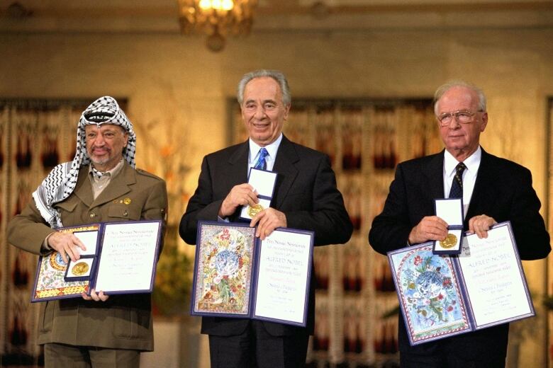 Three men stand in a line holding certificates in folder and small boxes containing round, gold medals.