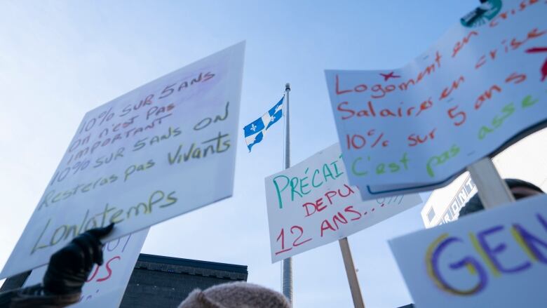 Signs at a protest hoisted in the air before a Quebec flag. 