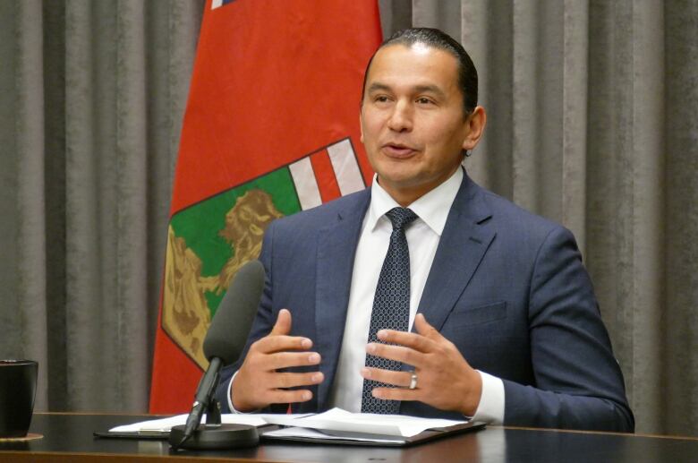 A man in a suit, seated at a table with a microphone and papers on it, speaks.