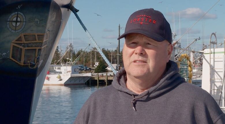 A white man wearing a grey hoodie and a black cap with 'Canadian Lobster' written on it stands on a harbour.