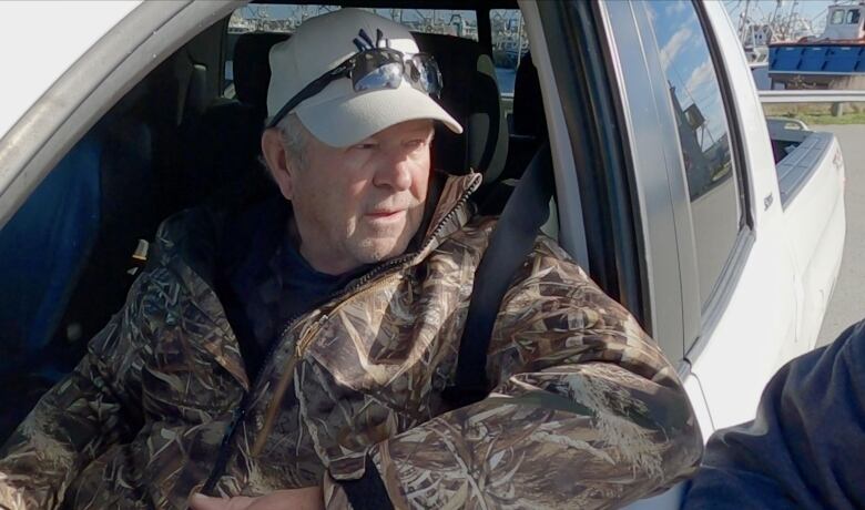 A white man in a camo jacket and a white cap sits inside a white truck.