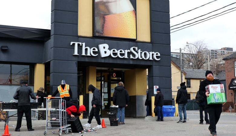 People are seen outside a Beer Store location, including one man carrying two cases of beer. 