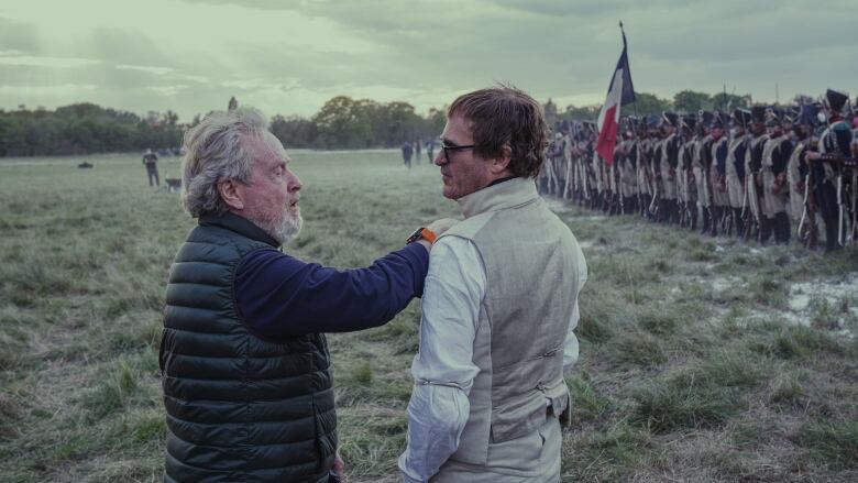 One man reaches over to adjust the costume of another. The man on the left wears modern clothes, while the man on the left is dressed in period clothes. Behind them an 18th century army stands on a field, holding a large French flag. 