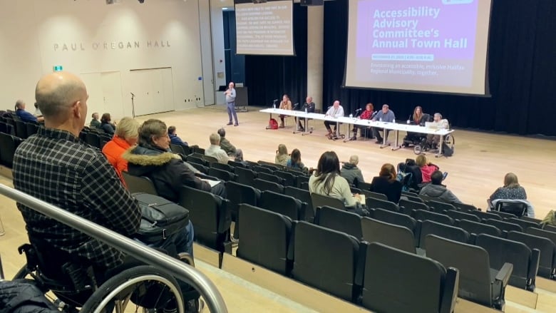 A view over an auditorium with people filling seats stepped down to a wide floor. A man in a wheelchair sits at the top row, with a panel of people at a table on the ground floor