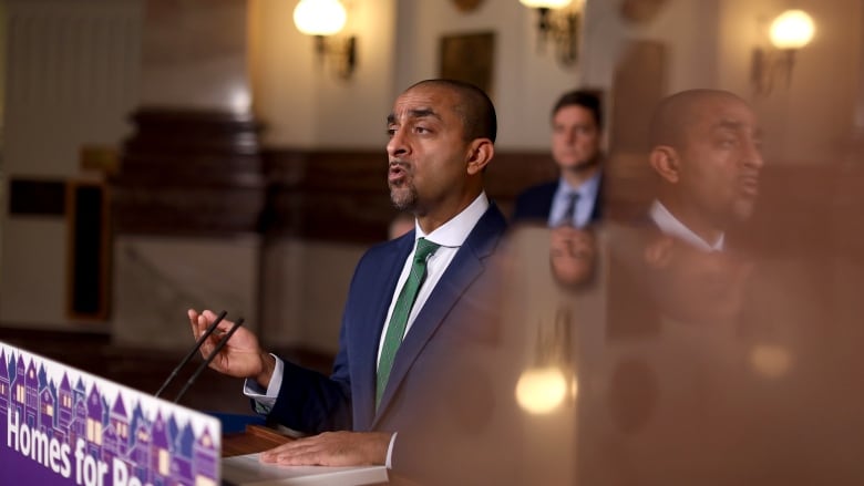 A South Asian man wearing a blue suit talks from a podium.