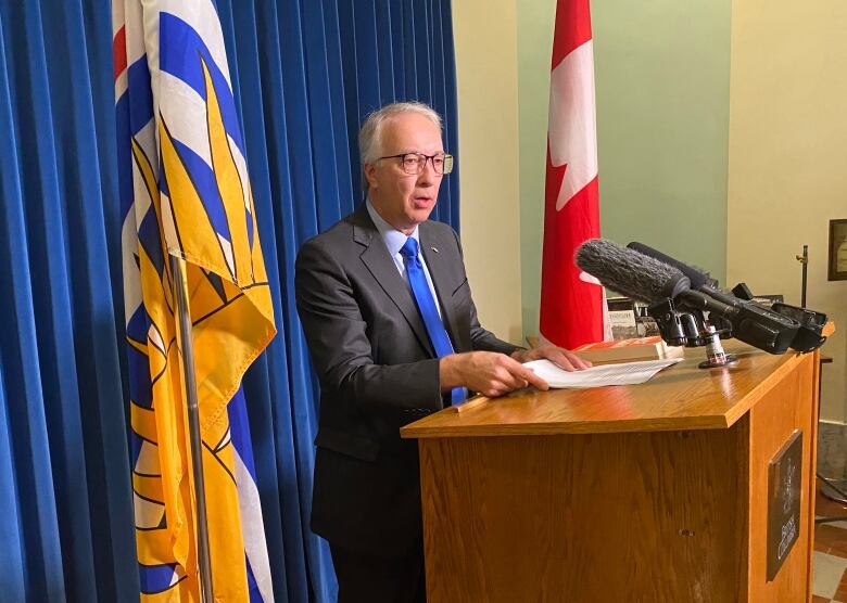 A white male with white hair wearing a black suit and blue tie stands at a podium.