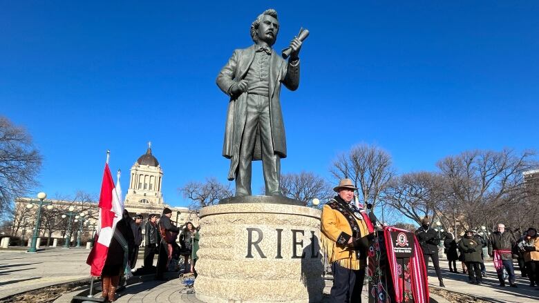 A man is shown speaking at a podium in front of a statue of a man, with a crowd surrounding them.