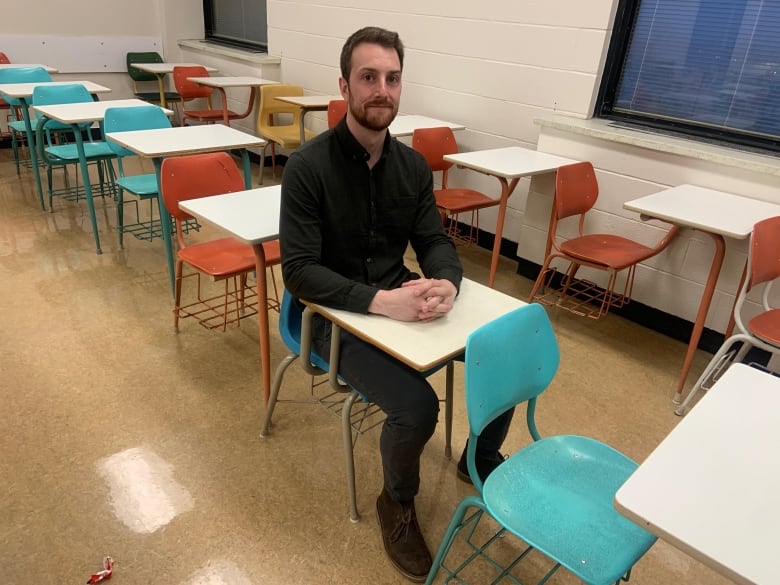 A man in a green button up shirt and jeans sits in a desk in a university classroom.