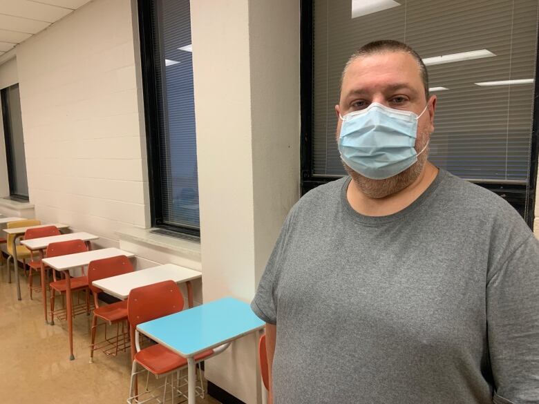 A man in a grey t-shirt stands in front of a window in a university classroom. 