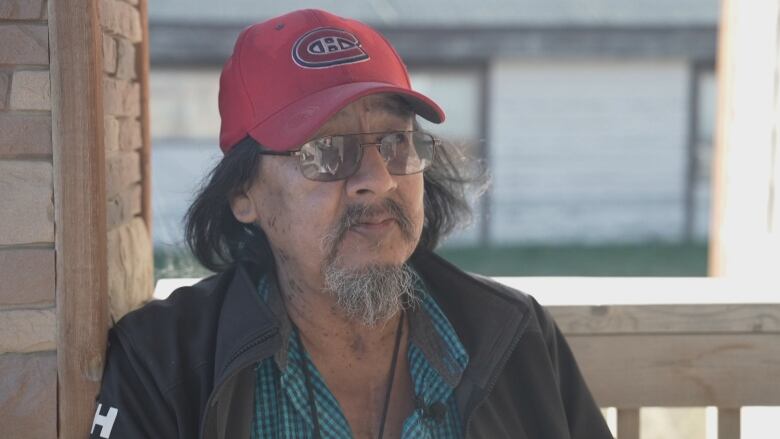 A man in a ball cap and glasses leans against a building.