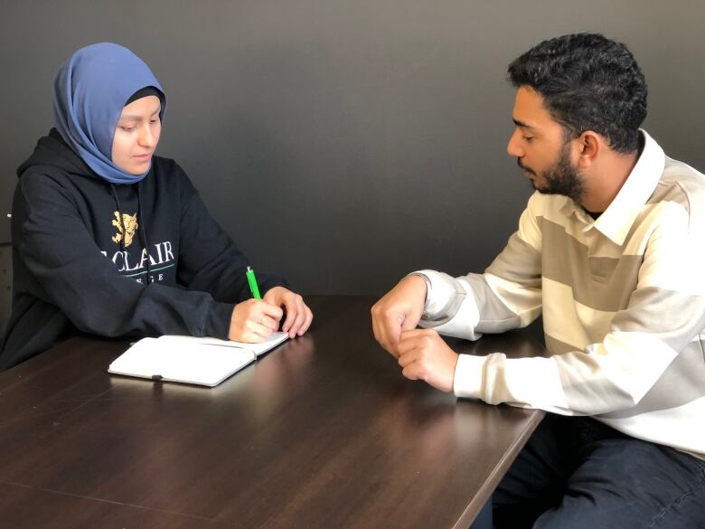 A woman wearing a black hoodie and a blue hijab sitting across from a man with black hair and facial hair wearing a white and grey-striped shirt